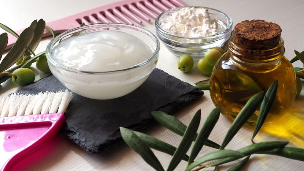 Afin d'illustrer l'article masque cheveux maison, des ustensiles et récipients sont posés sur une table. on voit 1 peigne, un pinceau, des feuilles d'oliviers, et des olives. Un petit bol translucide contient une matière qui semble visqueuse et est posé sur un gant de toilette gris foncé. Un récipient semble aussi contenir une matière couleur et matière miel.