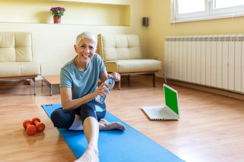 une femme âgée qui sourit assise sur un tapis bleu dans le cadre du sport pour personne âgée à la maison